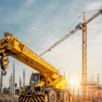 A construction site at dawn with a large yellow crane parked in front and scaffolding in the background.