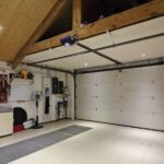 A garage interior with vaulted ceilings, recessed lighting, and tile floors. Shoes, outdoor tools, and firewood line the walls.