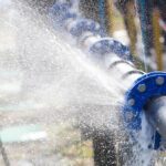 A burst metal pipe with blue connections with water spraying in streams out of it to the left and below.