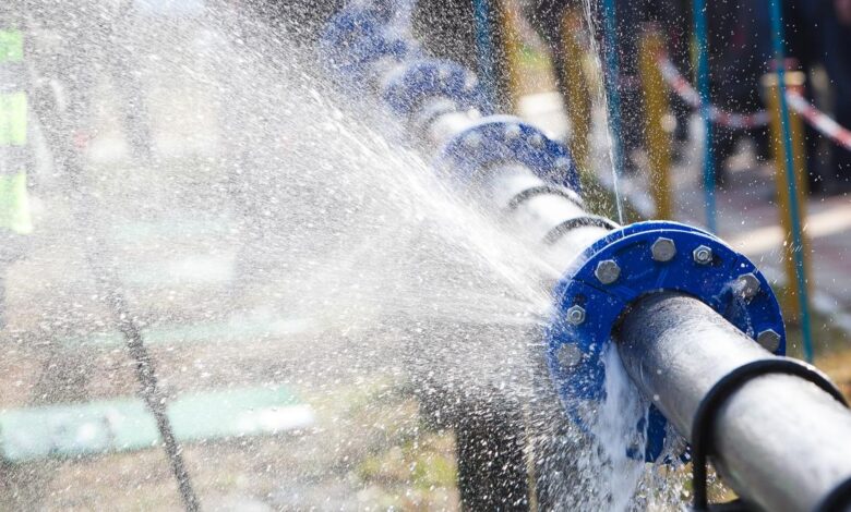 A burst metal pipe with blue connections with water spraying in streams out of it to the left and below.