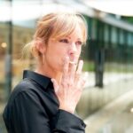 A blonde-haired woman wearing a black collared shirt smoking a cigarette while standing outside a building.