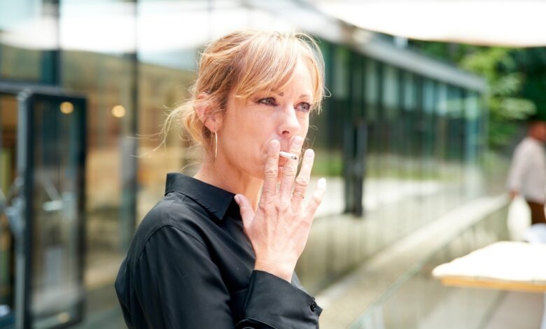 A blonde-haired woman wearing a black collared shirt smoking a cigarette while standing outside a building.
