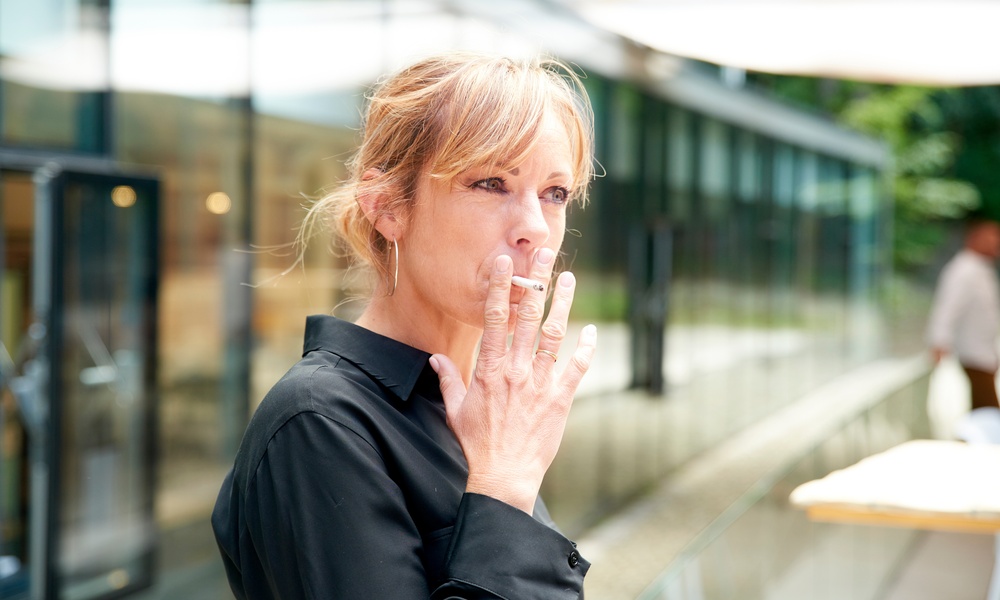 A blonde-haired woman wearing a black collared shirt smoking a cigarette while standing outside a building.