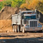A black and gray dump truck sits on a muddy worksite. A large pile of dirt sits behind the truck.