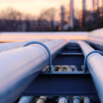 A close-up view shows four white industrial pipes hooked to a steel infrastructure. The sun sets in the background.