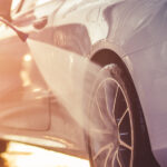 A close-up view shows a pressure washer washing the black tire of a white car. The sunshine shines under the car.