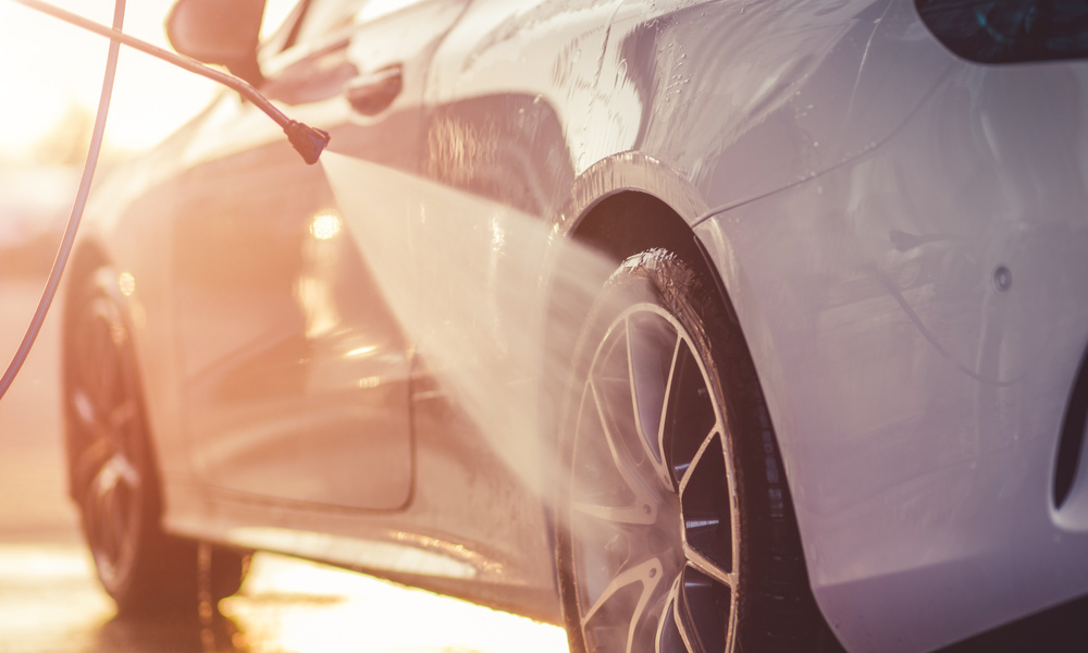 A close-up view shows a pressure washer washing the black tire of a white car. The sunshine shines under the car.