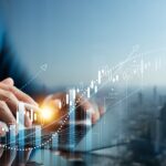 A person in a blue suit sitting at a table using a laptop with images representing financial growth superimposed on top.