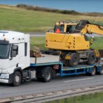A white truck on an open road with a drop deck trailer. A large yellow excavator crane is strapped to the trailer.