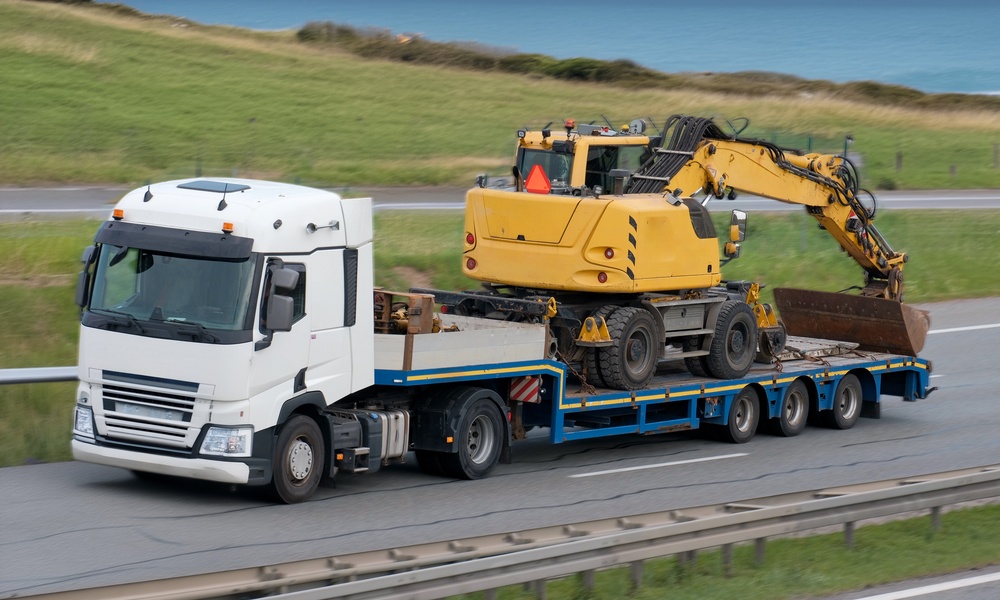 A white truck on an open road with a drop deck trailer. A large yellow excavator crane is strapped to the trailer.