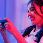 A smiling young person sitting in an office chair, wearing headphones, and holding up a gaming device.