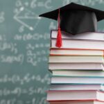 A stack of books sits on a desk in front of a chalkboard. A graduation cap sits on top of the books.