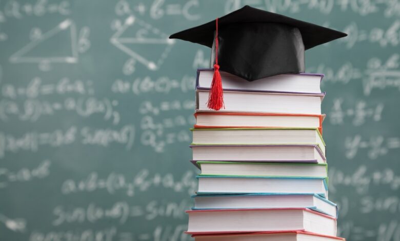 A stack of books sits on a desk in front of a chalkboard. A graduation cap sits on top of the books.