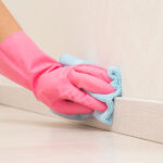 A close-up of someone cleaning their home's baseboards. They are wearing a pink glove and using a blue rag.