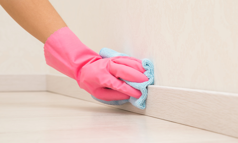 A close-up of someone cleaning their home's baseboards. They are wearing a pink glove and using a blue rag.