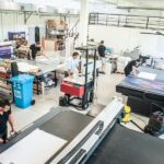 Five machine technicians work on various large-format printing machines in a commercial printing facility.