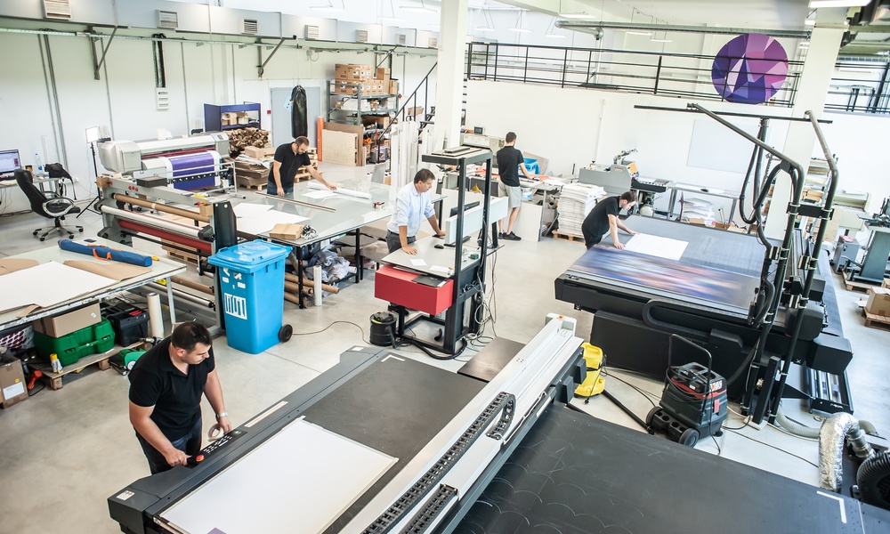 Five machine technicians work on various large-format printing machines in a commercial printing facility.