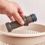 A person holds part of a plastic plumbing pipe in their left hand above a beige wastebasket near a wall.