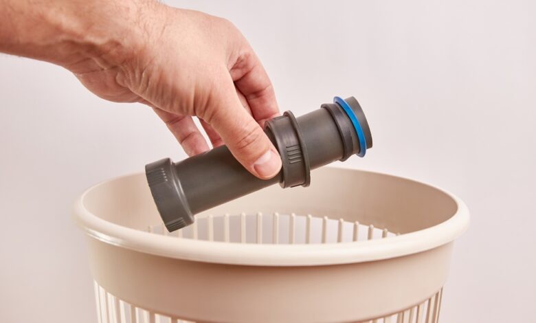 A person holds part of a plastic plumbing pipe in their left hand above a beige wastebasket near a wall.