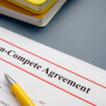 A document with a header reading "Non-Compete Agreement." A yellow pen rests on the paper, and glasses and notebooks sit nearby.