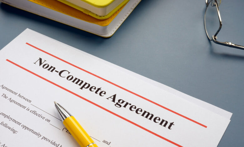 A document with a header reading "Non-Compete Agreement." A yellow pen rests on the paper, and glasses and notebooks sit nearby.
