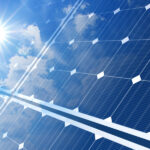 A close-up of a solar panel with multiple rows of small cells with a reflection of the sun and some clouds around it.
