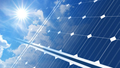 A close-up of a solar panel with multiple rows of small cells with a reflection of the sun and some clouds around it.