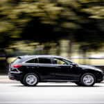 A black SUV contains a person in the driver's seat and driving down a suburban road surrounded by trees.