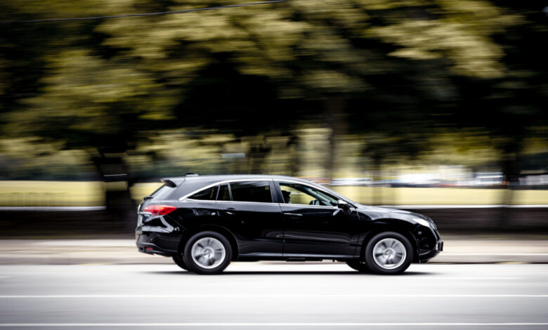 A black SUV contains a person in the driver's seat and driving down a suburban road surrounded by trees.