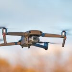 A silver drone quadcopter drone with a camera attached to the front of it flies outside.