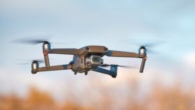 A silver drone quadcopter drone with a camera attached to the front of it flies outside.