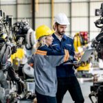 Two workers in a warehouse wear hard hats and look at a laptop and point at it while there's robotics equipment around them.