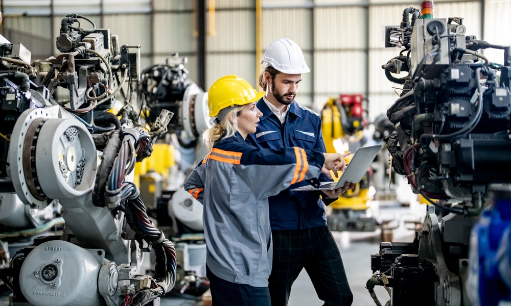 Two workers in a warehouse wear hard hats and look at a laptop and point at it while there's robotics equipment around them.