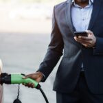 A business professional in their work suit holding their phone in one hand and charging their work car with the other hand.