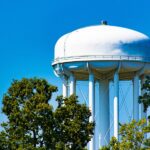 A white dome-shaped water tower against a clear blue sky. Several green trees are growing around it.