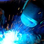 A person wearing a welding mask works on a project, with sparks flying, obscuring the details of the materials.