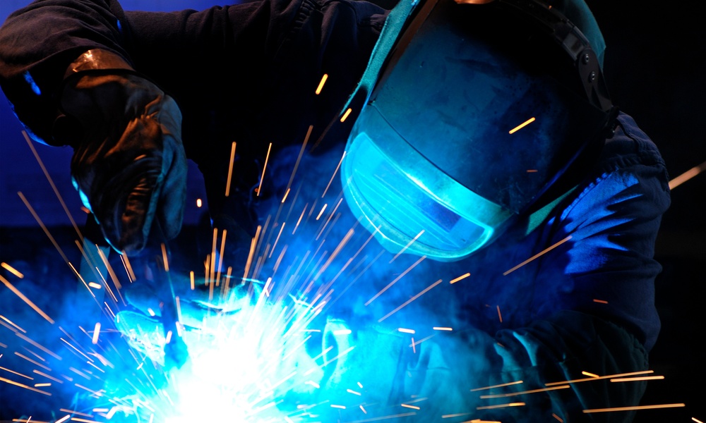 A person wearing a welding mask works on a project, with sparks flying, obscuring the details of the materials.