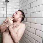 A man stands in a shower, crosses his arms over his chest, and looks up horrified at the running shower head.