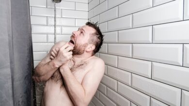 A man stands in a shower, crosses his arms over his chest, and looks up horrified at the running shower head.