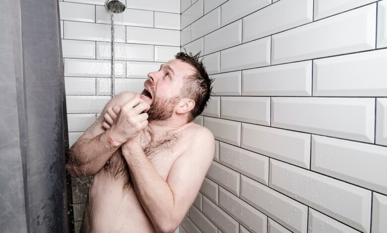 A man stands in a shower, crosses his arms over his chest, and looks up horrified at the running shower head.