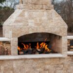 A large outdoor fireplace with a tall stone chimney burns multiple wood logs. A yard with trees is in the background.