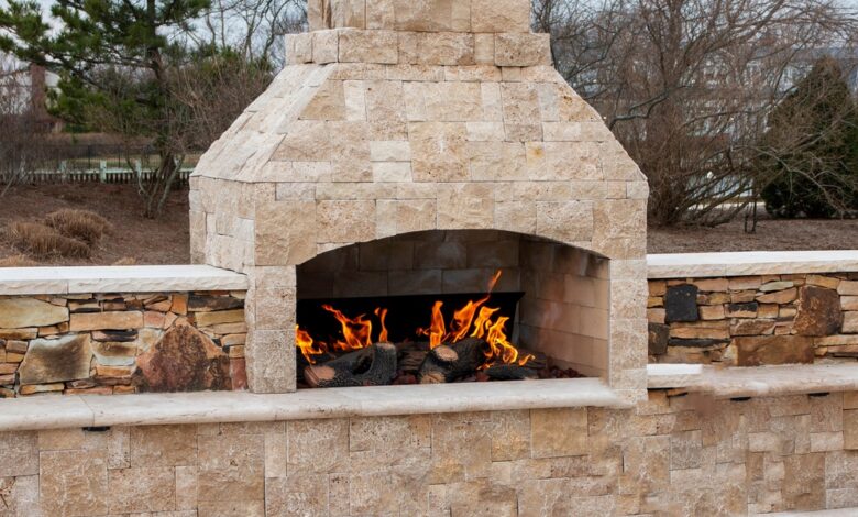 A large outdoor fireplace with a tall stone chimney burns multiple wood logs. A yard with trees is in the background.