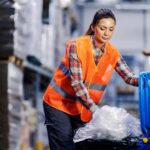 A female warehouse worker in an orange safety vest stuffs bundled plastic into a blue recycling bin.