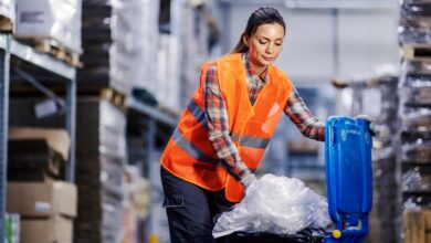 A female warehouse worker in an orange safety vest stuffs bundled plastic into a blue recycling bin.