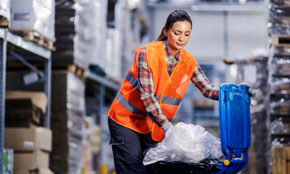 A female warehouse worker in an orange safety vest stuffs bundled plastic into a blue recycling bin.
