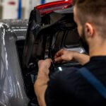 An auto mechanic reworking the tires in a car's trunk lid. The trunk is open and they're working on lighting wires.