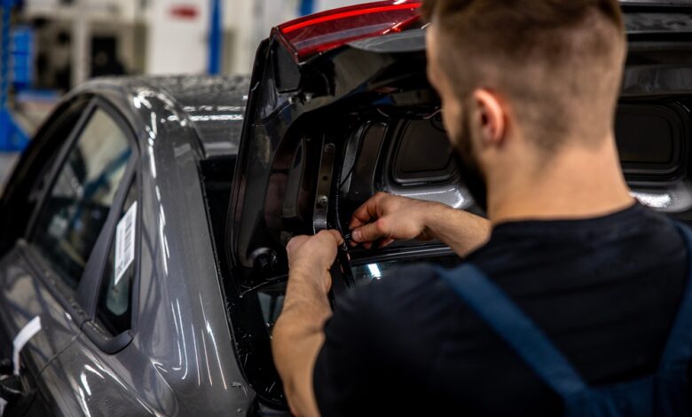 An auto mechanic reworking the tires in a car's trunk lid. The trunk is open and they're working on lighting wires.