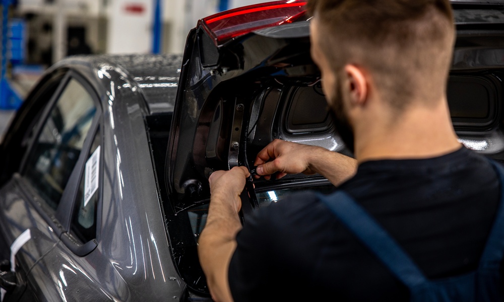 An auto mechanic reworking the tires in a car's trunk lid. The trunk is open and they're working on lighting wires.
