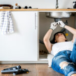 A woman wearing ripped jeans and a white t-shirt lays under her kitchen sink to repair it.