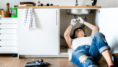 A woman wearing ripped jeans and a white t-shirt lays under her kitchen sink to repair it.